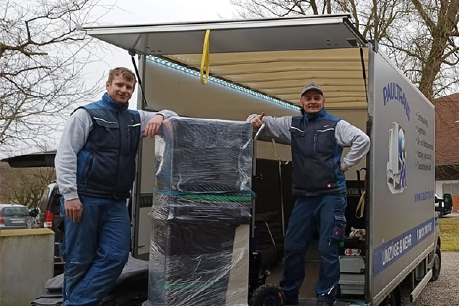 zwei Umzugsmitarbeiter stehen neben verpacktem Möbelstück vor offenem Umzugswagen