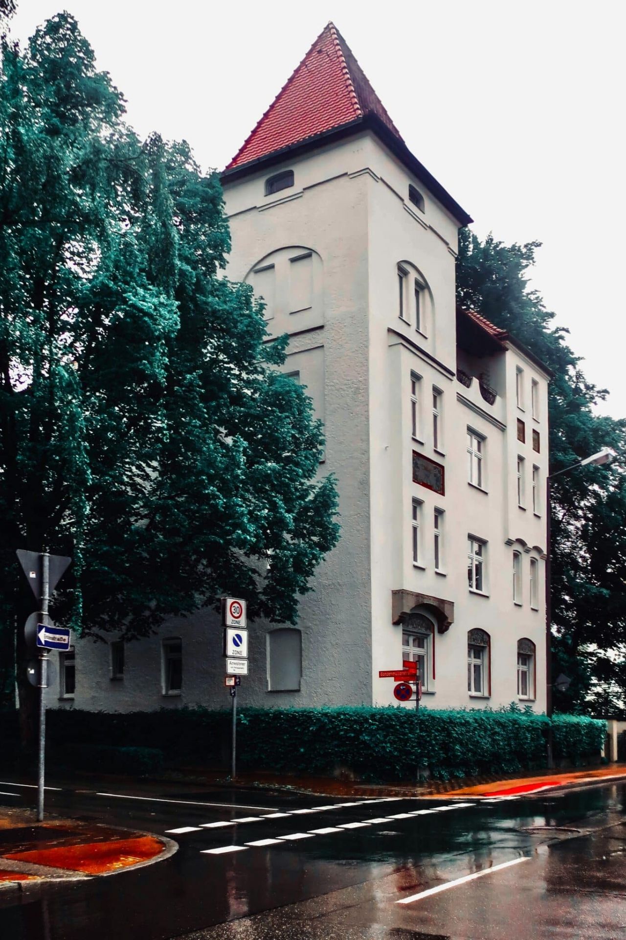 Altbau mit Turm und rotem Dach an einer nassen Straßenecke, umgeben von Bäumen